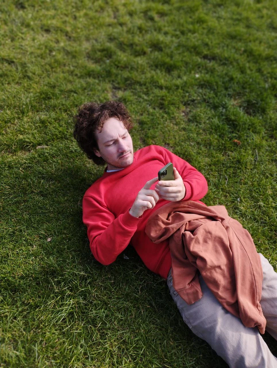 A person in a red sweater lying in the grass and looking at a phone.